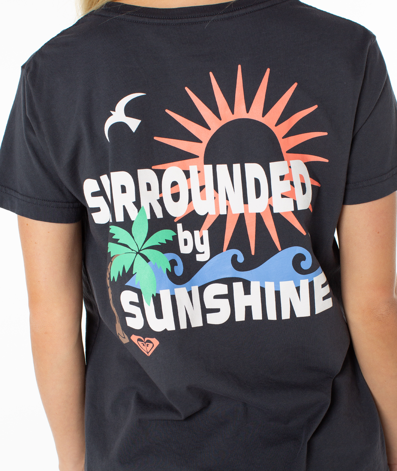 A person wearing the Roxy Surrounded By Sunshine Tee, a black slouchy T-shirt featuring "Surrounded by Sunshine" with sun, palm tree, bird, and blue waves graphics on the back.
