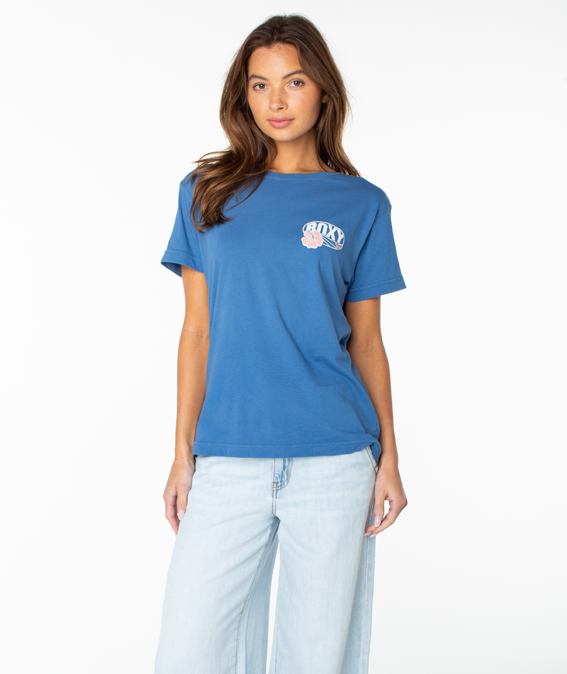 A woman with long brown hair stands against a plain white background, wearing the Roxy OG Roxy Oval Boyfriend Crew in Dutch Blue Og Roxy Oval paired with light blue jeans.