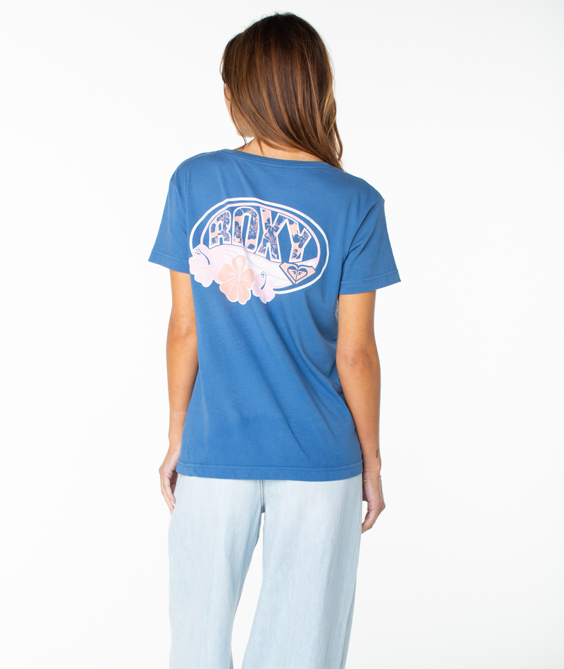 A woman with long brown hair wears the Roxy OG Roxy Oval Boyfriend Crew, a blue crewneck top featuring pink floral graphics on the back, paired with light blue jeans, and stands against a white background.