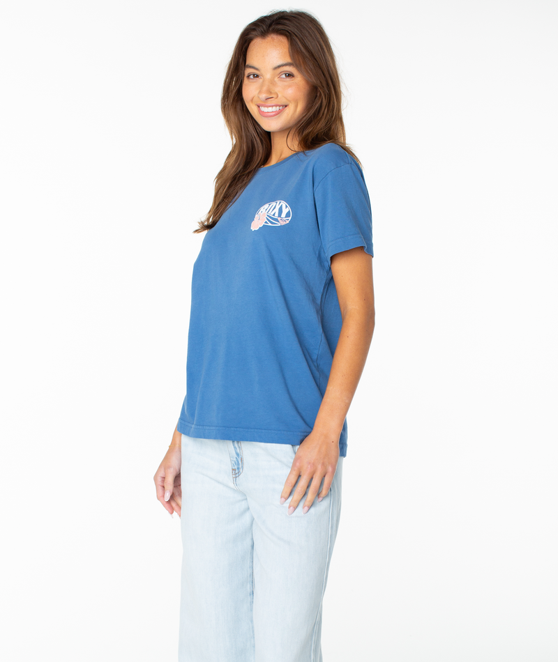 A woman with long brown hair is wearing the OG Roxy Oval Boyfriend Crew by Roxy—a blue beachy top featuring a small shell design on the chest—paired with light blue jeans, smiling and standing against a plain white background.