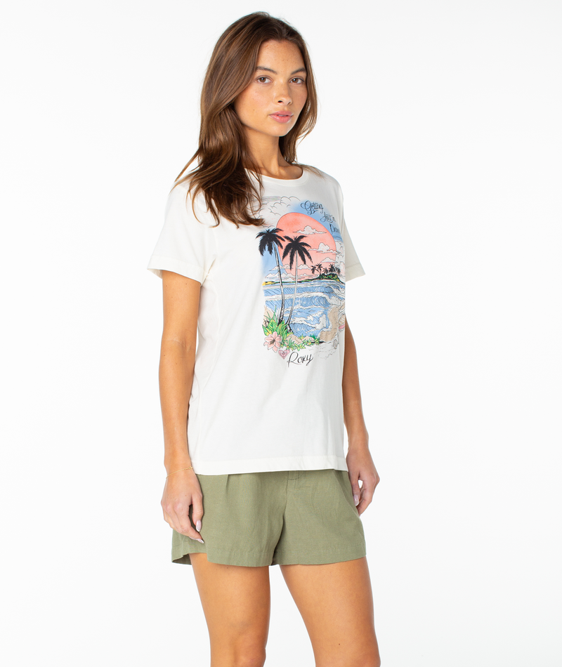 A woman with long brown hair wears the Roxy Coastal Beach Boyfriend Crew, a slouchy white graphic t-shirt with a tropical beach design, paired with green shorts and standing against a plain white background.