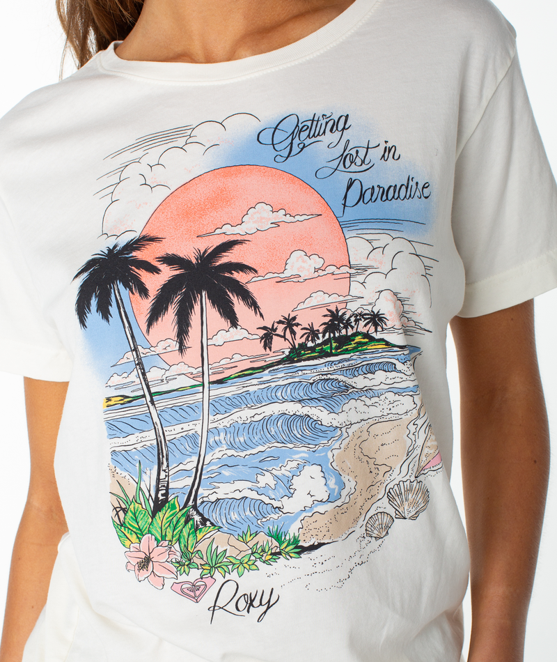 A woman wears the Roxy Coastal Beach Boyfriend Crew, a slouchy top featuring a beach scene with palm trees, waves, seashells, and a pink sun. The text reads “Getting Lost in Paradise” and “Roxy.”.