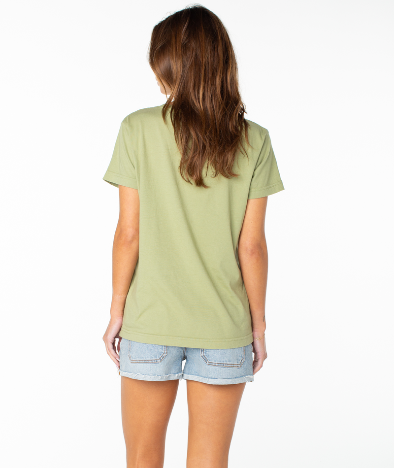 A woman with long brown hair faces away from the camera, wearing the Roxy Shell Band Boyfriend Crew in light green and denim shorts against a white background.