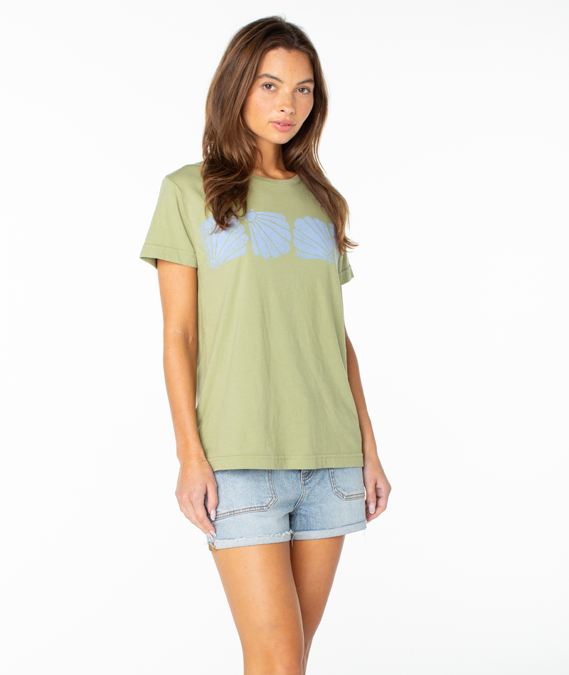 A woman wearing the Roxy Shell Band Boyfriend Crew—a light green beachy top with blue shell designs—paired with denim shorts stands against a white background.