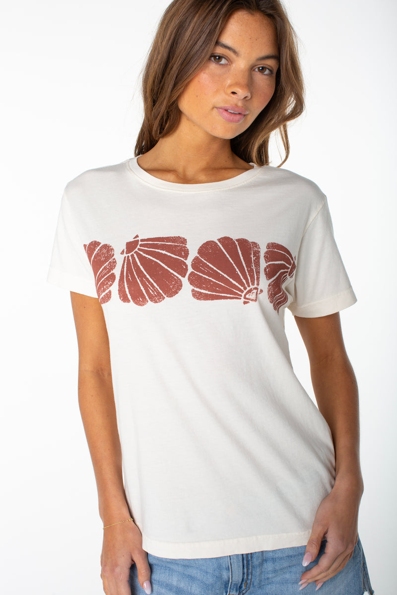 A woman stands against a plain white background wearing the Roxy Shell Band Boyfriend Crew, a slouchy off-white t-shirt featuring red shell prints across the chest.