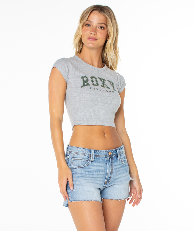 A woman wears a grey Roxy Academy Tee with green text across the chest and light-wash denim shorts.