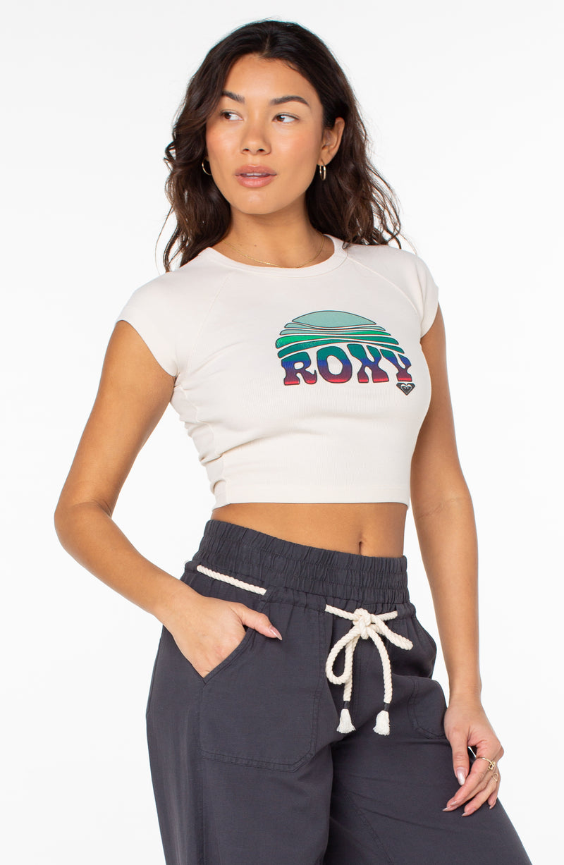 A cropped, cream-colored Roxy Rising Tee with cap sleeves and a graphic of a sun setting over the ocean, worn with dark drawstring pants.