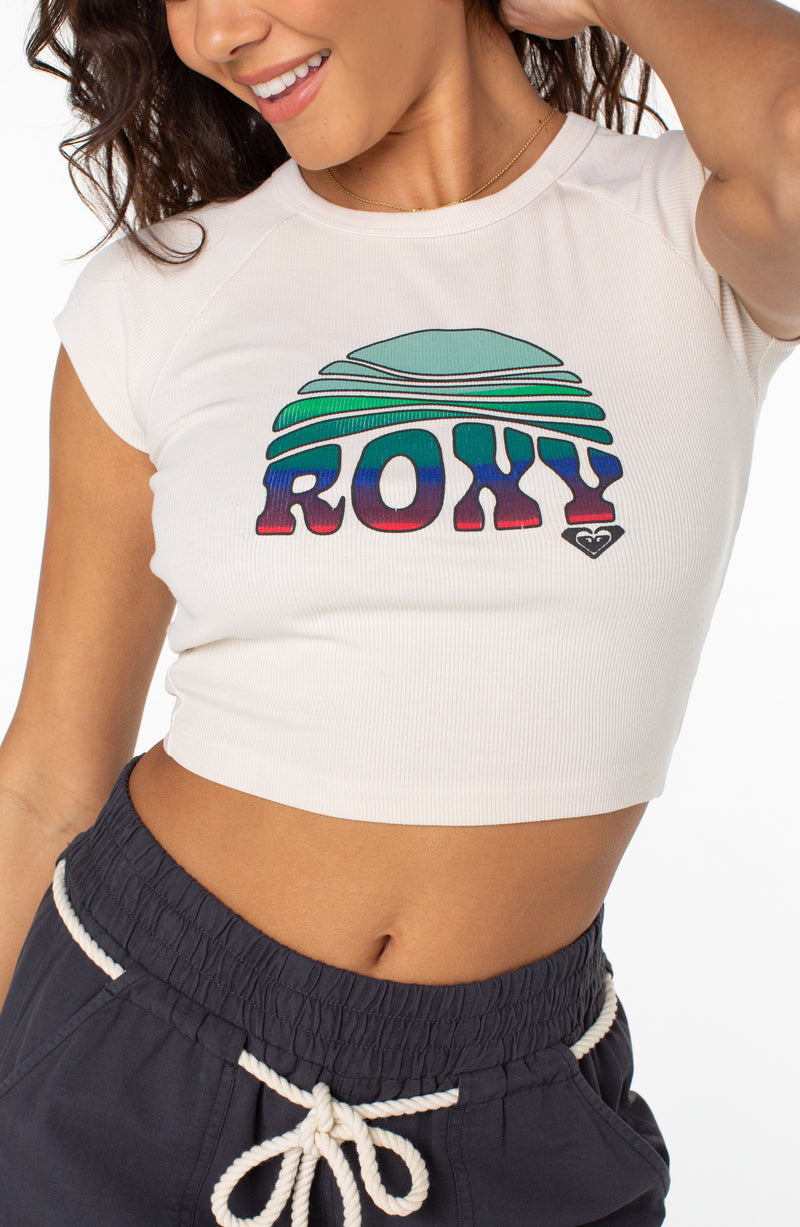 A woman wears a white, ribbed, cropped Roxy Rising Tee with cap sleeves and a graphic of a sun setting over the ocean on the chest.