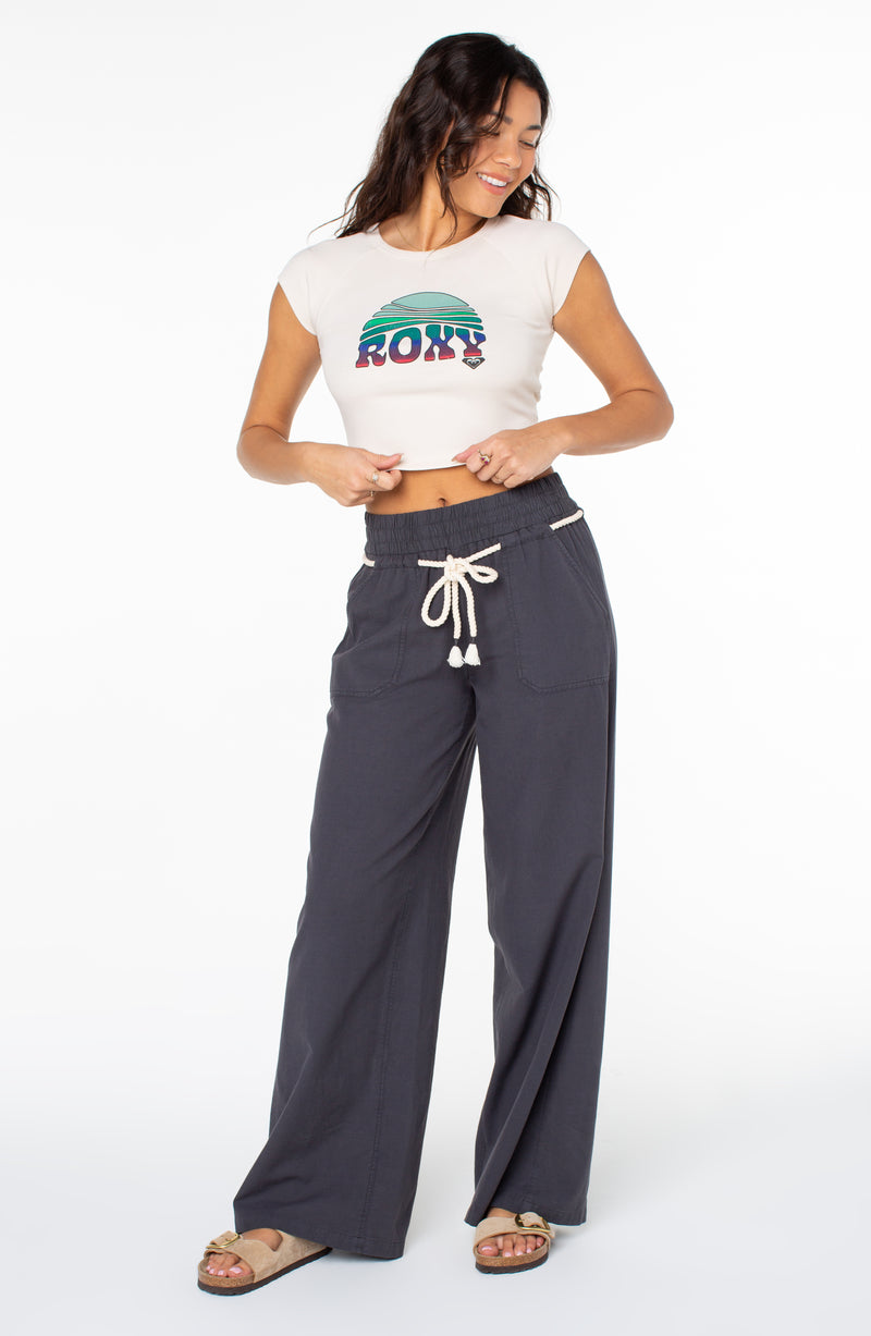 A person wearing a cream-colored Roxy Rising Tee with a colorful graphic and dark gray wide-leg pants with a white drawstring waist.