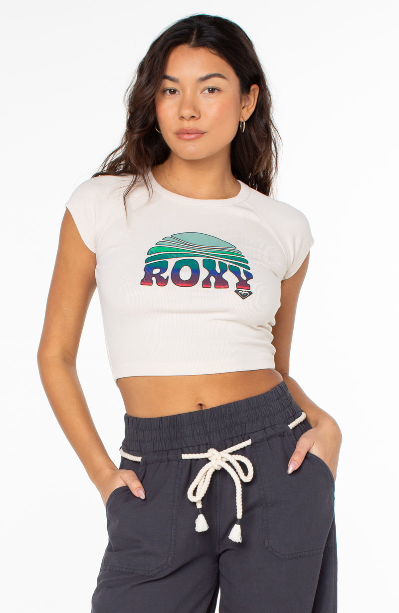 A person wears a cream-colored, cropped cap-sleeve Roxy Rising Tee with a vintage-style graphic of a sun over mountains on the chest.
