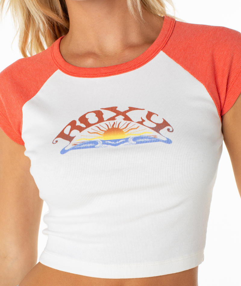 A person wears the Garrett Wave Tee by Roxy, a white and orange graphic crop top with “Roxy” and a rising sun over waves—perfect as an effortless spring staple for casual outfits.