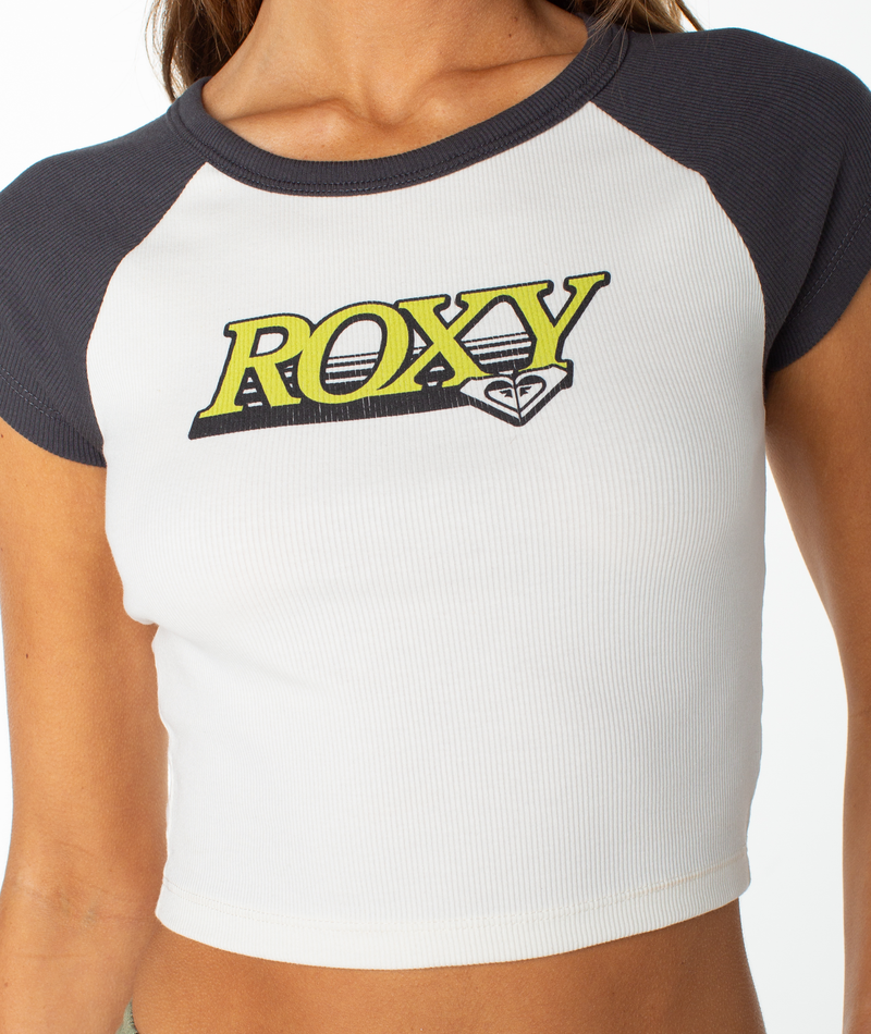 A person wearing the Roxy Crop Baby Tee, a short-sleeve white top with dark sleeves and a yellow ROXY logo on the chest by Roxy—perfect for adding a graphic touch to any capsule wardrobe.