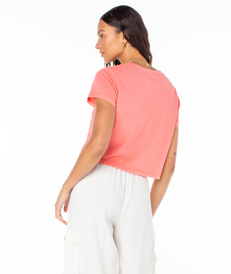 A person viewed from behind wearing a coral pink Roxy Paraiso Screen Tee and white trousers.