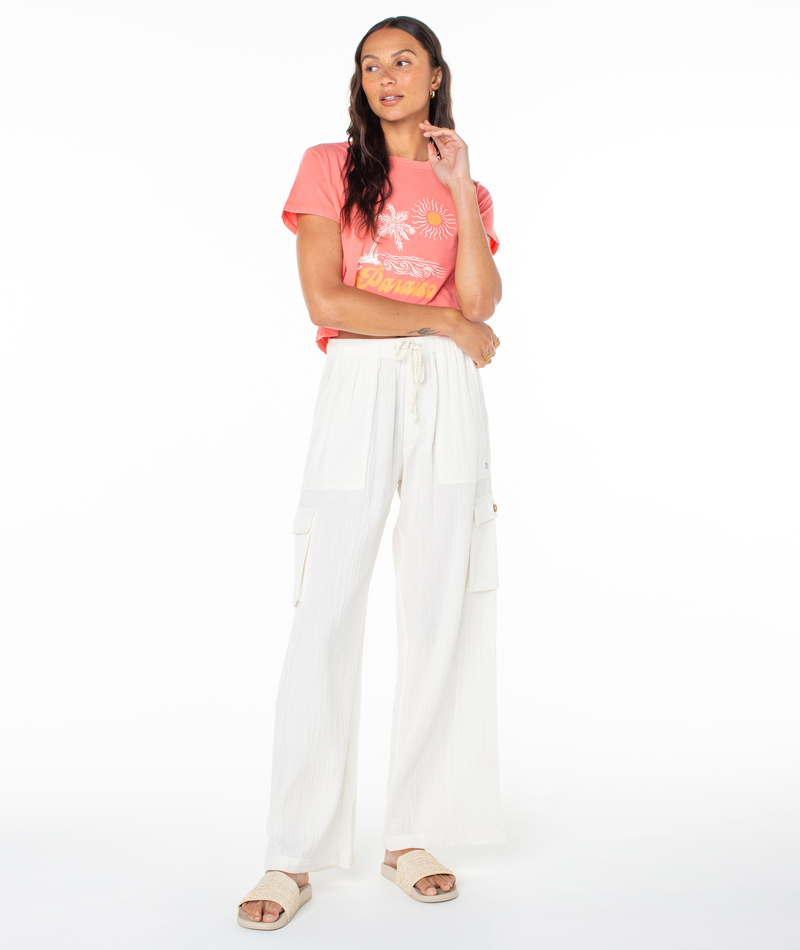 A woman wears a Roxy Paraiso Screen Tee in coral with a tropical graphic and white wide-leg cargo pants with a drawstring waist.