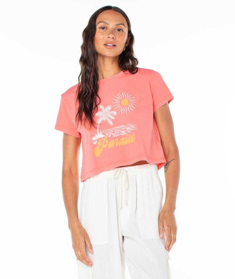 A woman wears a Roxy Paraiso Screen Tee, a coral pink cropped t-shirt featuring a white and yellow graphic of a palm tree, sun, and the word Paraiso.