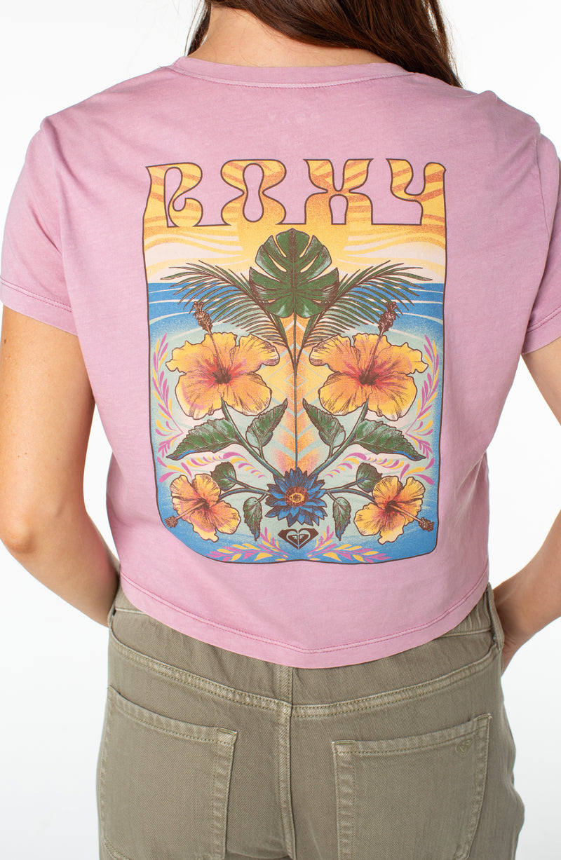 A person wears a dusty pink Roxy Slow Dive Tee featuring a back graphic of yellow hibiscus flowers, palm leaves, and a sunset landscape.