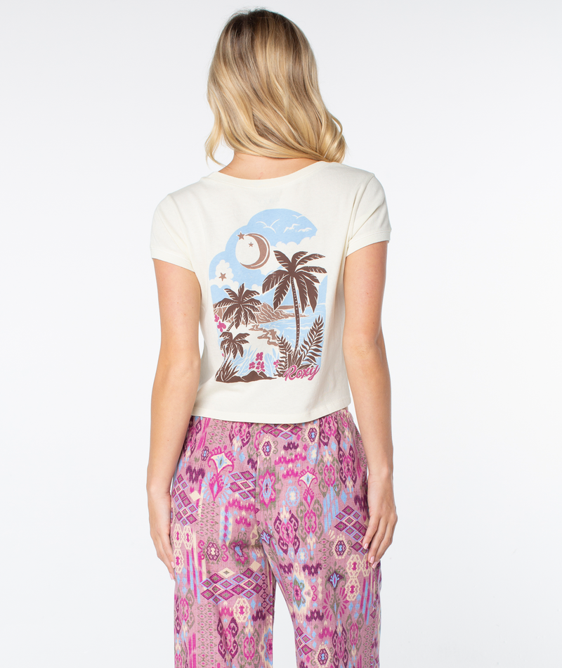 A woman wears a cream-colored Roxy Moon Beach Top featuring a graphic of a tropical beach scene, a crescent moon, and stars on the back, paired with patterned pink pants for a breezy fit.