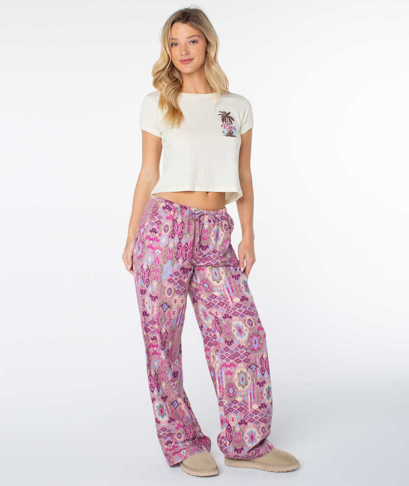 A woman wearing a Roxy Moon Beach Top in Cloud Dancer, a breezy, cream-colored cropped T-shirt with a small coastal cool palm tree graphic, paired with loose, patterned lounge pants in shades of pink, purple, and blue.