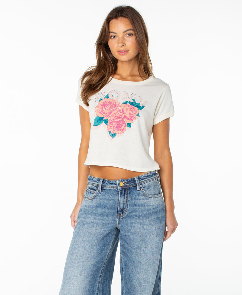 A woman wears the Roxy Rose Heart Cropped Baby Tee in Cloud Dancer Rose Heart, featuring a playful pink floral graphic, paired with blue jeans and standing against a plain white background—a true wardrobe staple.