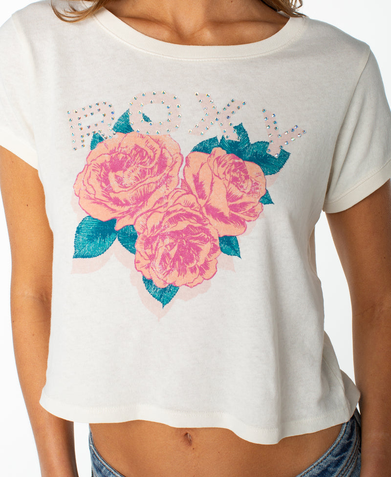Wearing the Rose Heart Cropped Baby Tee by Roxy, a woman sports this white top featuring ROXY in rhinestones above three large pink roses and green leaves printed on the front—a breezy fit with a playful graphic that makes it a true wardrobe staple.