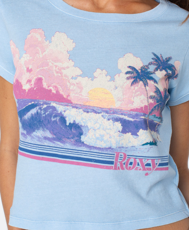 Wearing the Roxy Salt Sunrise Cropped Baby Tee in light blue, featuring a graphic of a pastel sunset with ocean waves, clouds, and palm trees in pink, purple, and yellow—a breezy essential for your wardrobe.