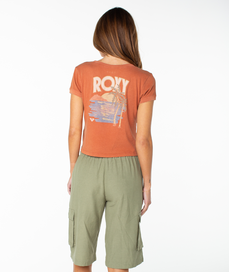 A woman faces away in a rust-colored Roxy Spanish Summer Cropped Baby Tee featuring a beach scene graphic, paired with green cargo shorts against a white background—a breezy fit sure to become a wardrobe staple.