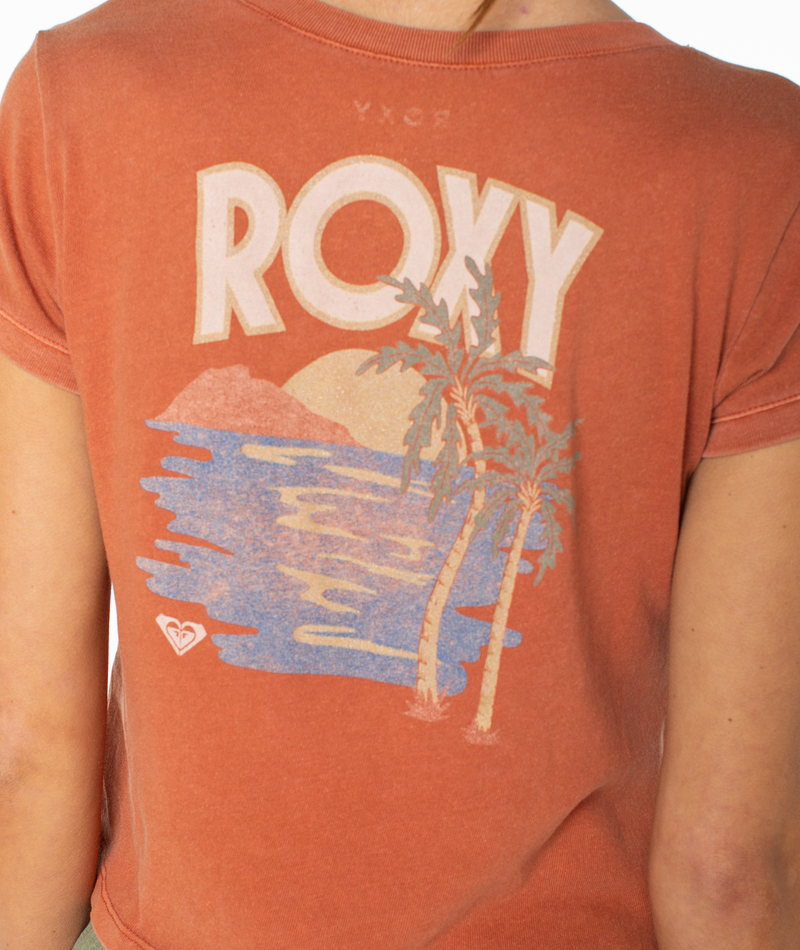 A person is seen from behind wearing the Roxy Spanish Summer Cropped Baby Tee—a breezy, faded orange graphic top featuring “ROXY” above an island scene with a sunset, water, palm trees, and mountains. The relaxed fit and stylish print make this tee a must-have wardrobe staple.