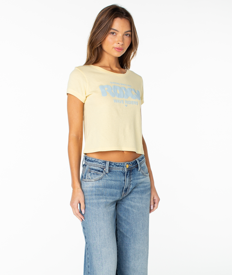 A woman with long brown hair wears the Roxy Wave Riders Club Cropped Baby Tee—a light yellow top with a blue logo—paired with high-waisted blue jeans for a breezy, staple look, standing against a plain white background.