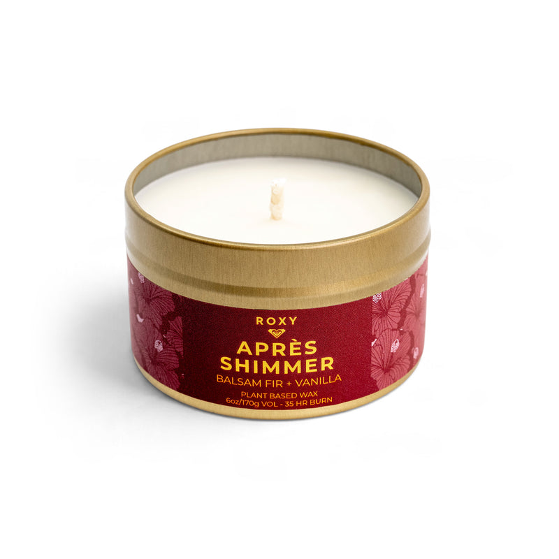 A ROXY Apres Shimmer Holiday Tin Candle in a gold-rimmed tin, featuring a red label with floral patterns and text reading Après Shimmer, Balsam Fir + Vanilla, evoking winter luxury.