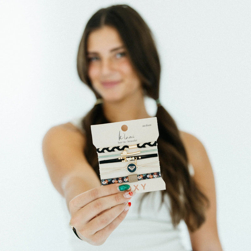 A woman with long brown hair in pigtails holds out a Roxy K'lani x Roxy Hair Tie Bracelet, its braided textures in focus as she smiles softly, her face slightly blurred in the background.