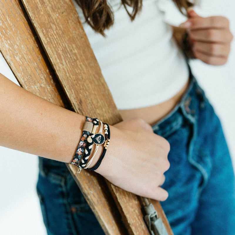 A person wearing a white crop top and blue jeans holds a wooden ladder. Their wrist features several surf-inspired accessories, including the K'lani x Roxy Hair Tie Bracelet by Roxy.