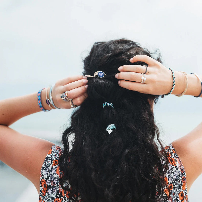 A person with long, wavy dark hair, seen from behind, adjusts their hair with both hands. They're wearing K'lani x Roxy Hair Tie Bracelets and rings by Roxy, plus a patterned sleeveless top from the Ocean Bloom set.