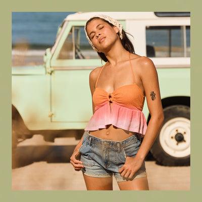 Woman in a colorful top and denim shorts standing in front of a vintage van on a beach.