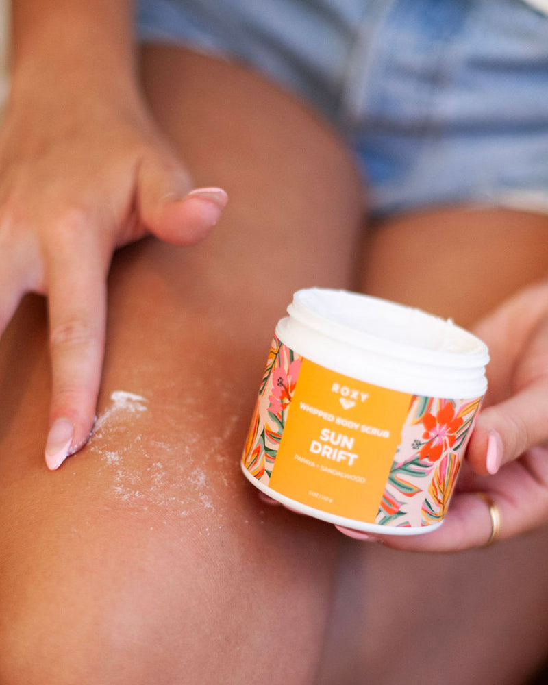 A person in denim shorts applies ROXY’s Sun Drift Body Butter & Scrub Bundle, featuring a white whipped formula with an orange floral design, to their leg—unveiling tropical notes that exfoliate, hydrate, and leave skin silky-soft.