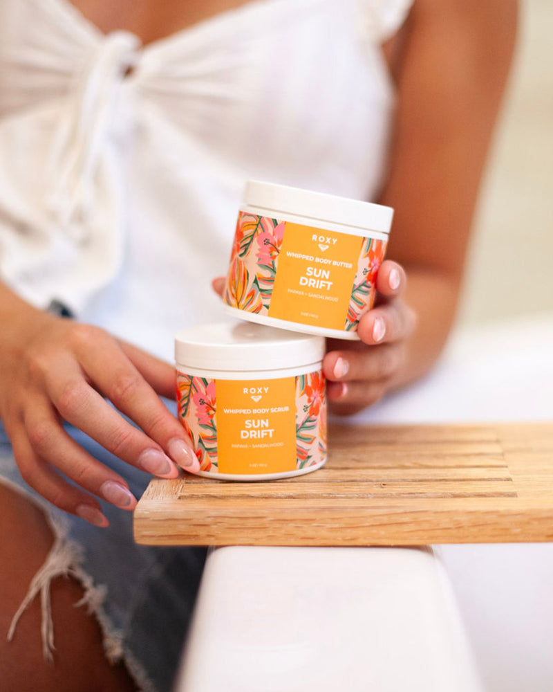 A person in a white top and denim shorts holds the ROXY Sun Drift Body Butter & Scrub Bundle, featuring two vibrant orange jars with colorful floral designs, on a wooden tray—ideal for anyone looking to exfoliate and hydrate for silky-soft skin with tropical notes.