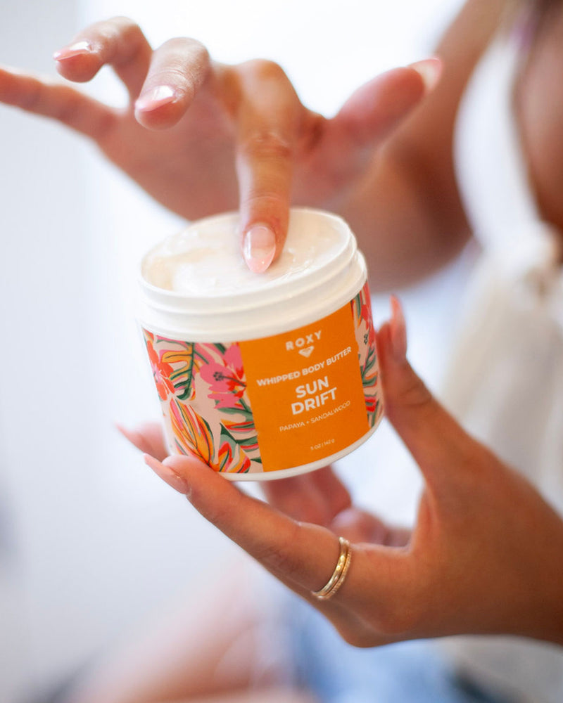 A person with manicured nails dips a finger into a jar from the ROXY Sun Drift Body Butter & Scrub Bundle, featuring a colorful floral label, to exfoliate and hydrate their skin for a silky-soft feel infused with tropical notes.