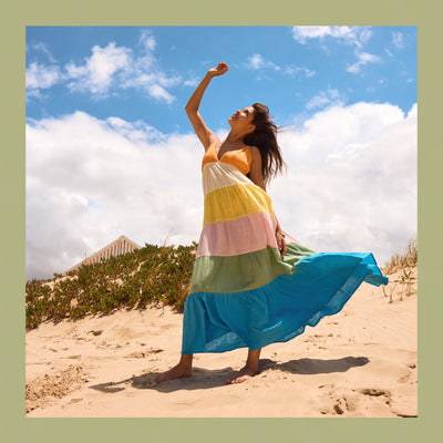 Woman in a colorful dress standing on a sandy beach with a blue sky and clouds in the background.