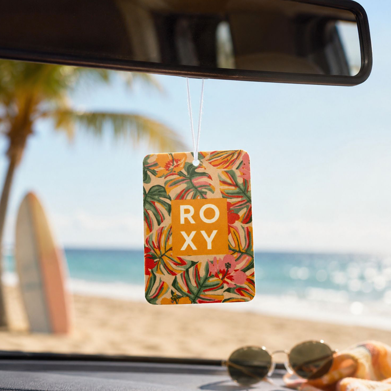 The Roxy Sun Drift Palm Air Freshener hangs from the rearview mirror, its sunlit scent filling the car. Outside, palm trees and beach waves beckon, while sunglasses on the dashboard add to the carefree coastal vibe.