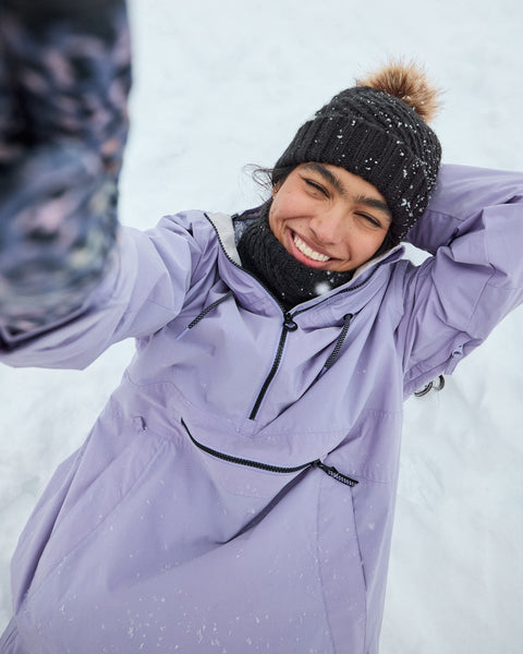 Wearing the Roxy Downtown Anorak, a person in a vibrant purple snow jacket and black knit hat smiles while taking a selfie in the snow, with snowflakes dusting their clothing and hat—highlighting both the coziness and style of this quality technical outerwear.