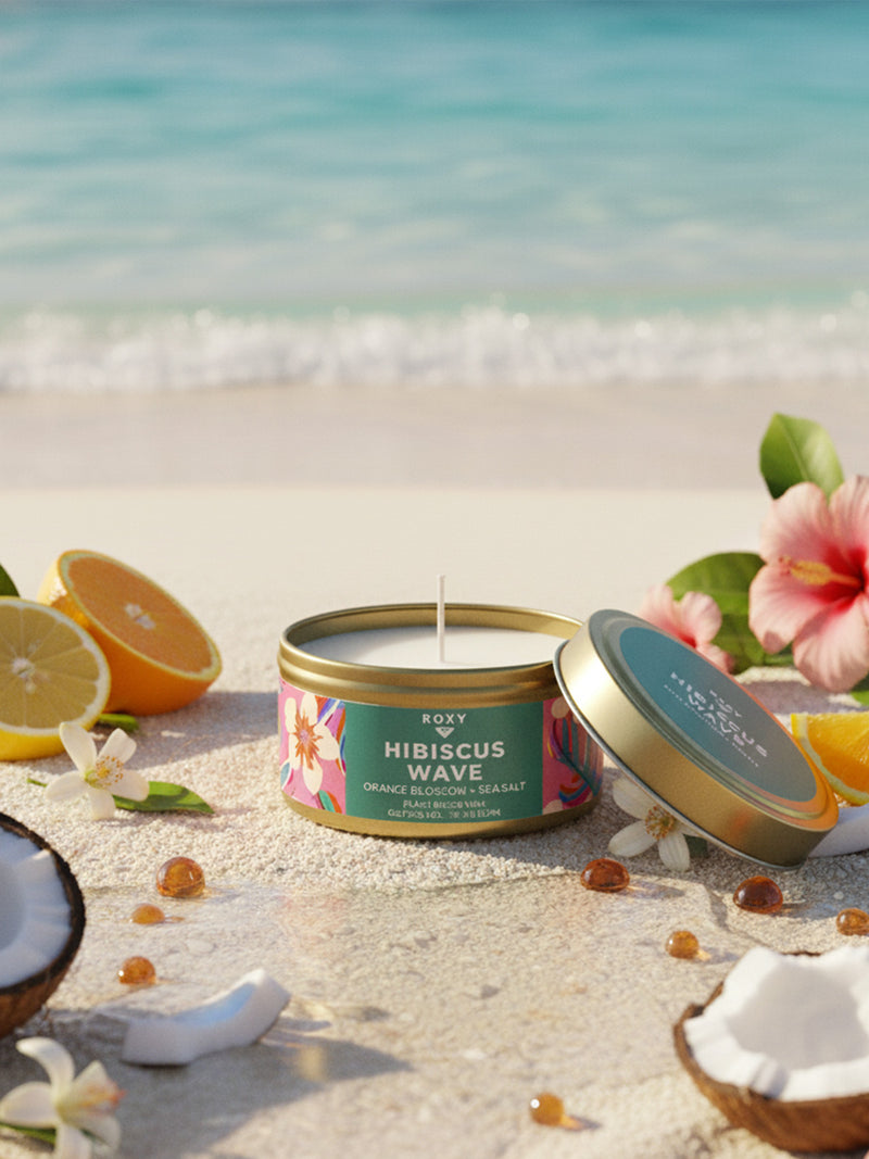 A Roxy Hibiscus Wave Palm Tin Candle with a tropical aroma sits open on a sandy beach, surrounded by coconut halves, orange blossom, hibiscus flowers, and amber stones, against a turquoise ocean and blue sky.