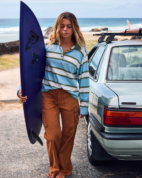 A young woman stands by a car holding a surfboard, wearing the Roxy A La Cargo Pant in an orange wash and a striped long-sleeve shirt, with the ocean and rocks visible in the background.