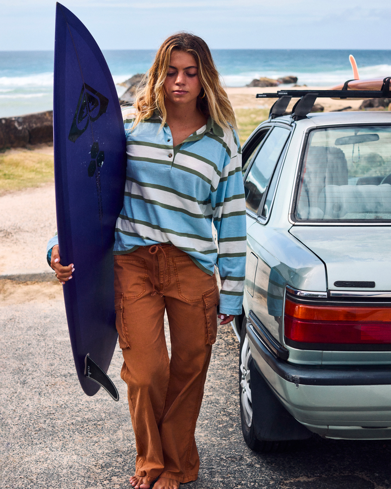 A young woman stands by a car holding a surfboard, wearing the Roxy A La Cargo Pant in an orange wash and a striped long-sleeve shirt, with the ocean and rocks visible in the background.