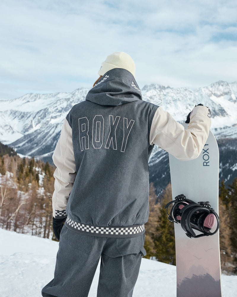 Wearing the Roxy Cheer Up Jacket, a person stands on snowy ground holding a snowboard and gazing at snow-covered mountains. The back of their women’s snow jacket displays the ROXY logo, with pine trees visible beneath the mountains.