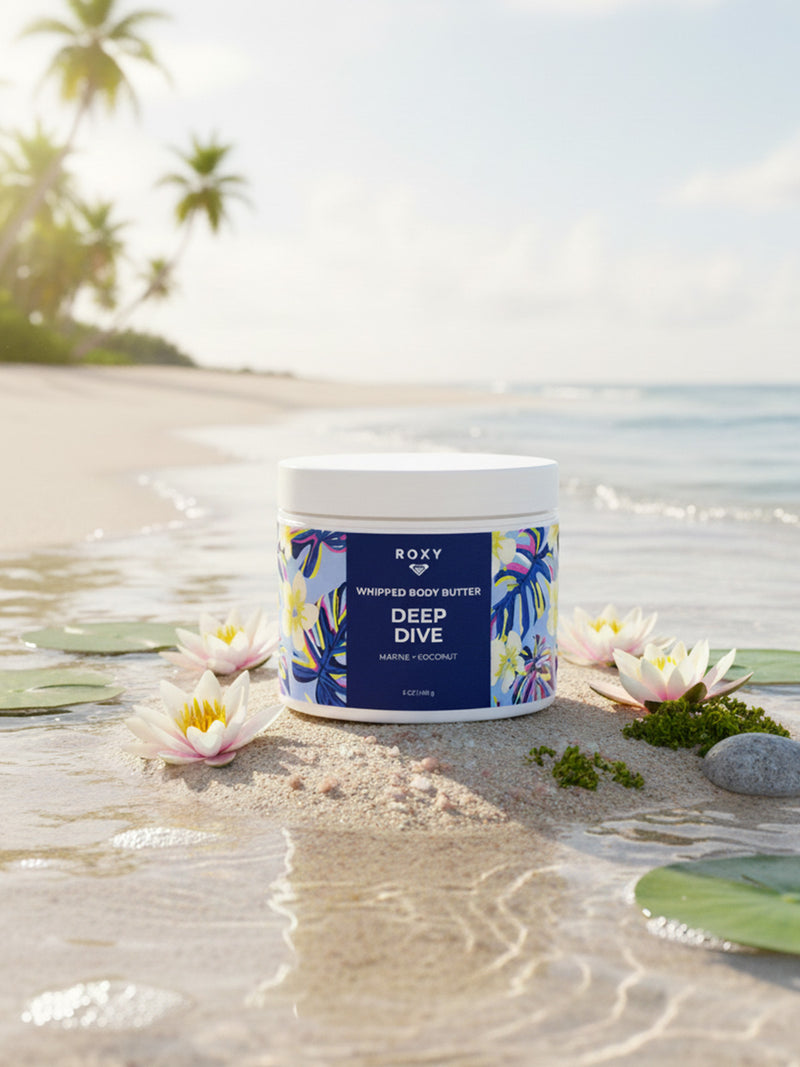 A jar of Roxy Deep Dive Body Butter rests on sand with tropical flowers and a palm leaf nearby. Behind it, a sunny beach, ocean waves, and palm trees evoke the marine notes and ocean-air softness of the product.