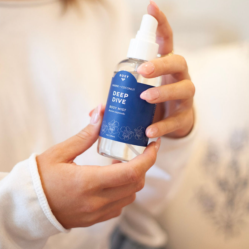 A person in a white long-sleeve shirt holds a clear Roxy Deep Dive Body Mist with a blue label and white cap, ready to spray the ocean-inspired scent enhanced with pink sea salt.