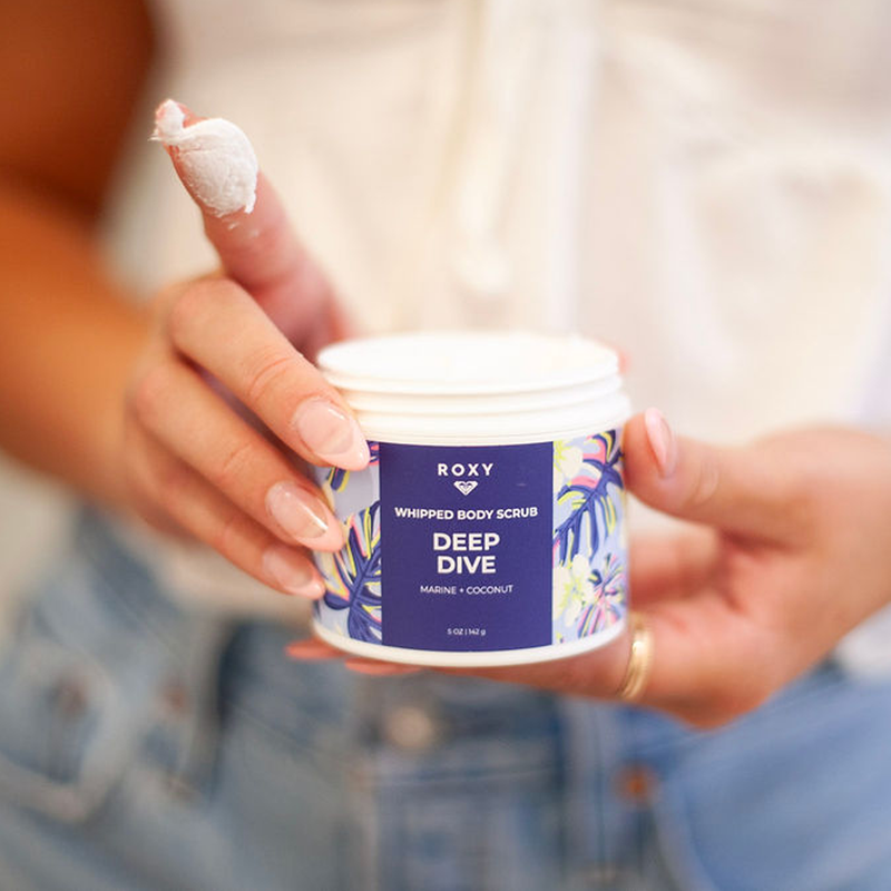 Someone holds an open jar of Roxy Deep Dive Body Scrub, with a bit on their finger. The blue floral label pops against their white top and jeans, making this scrub the perfect way to upgrade any shower routine.