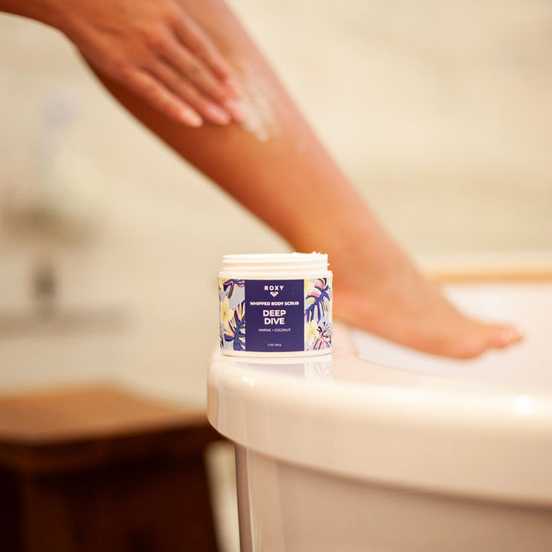 A person applies lotion to their leg as a jar of Roxy Deep Dive Body Scrub rests on the bathtub’s edge, the softly blurred background creating a mindful self-care moment.