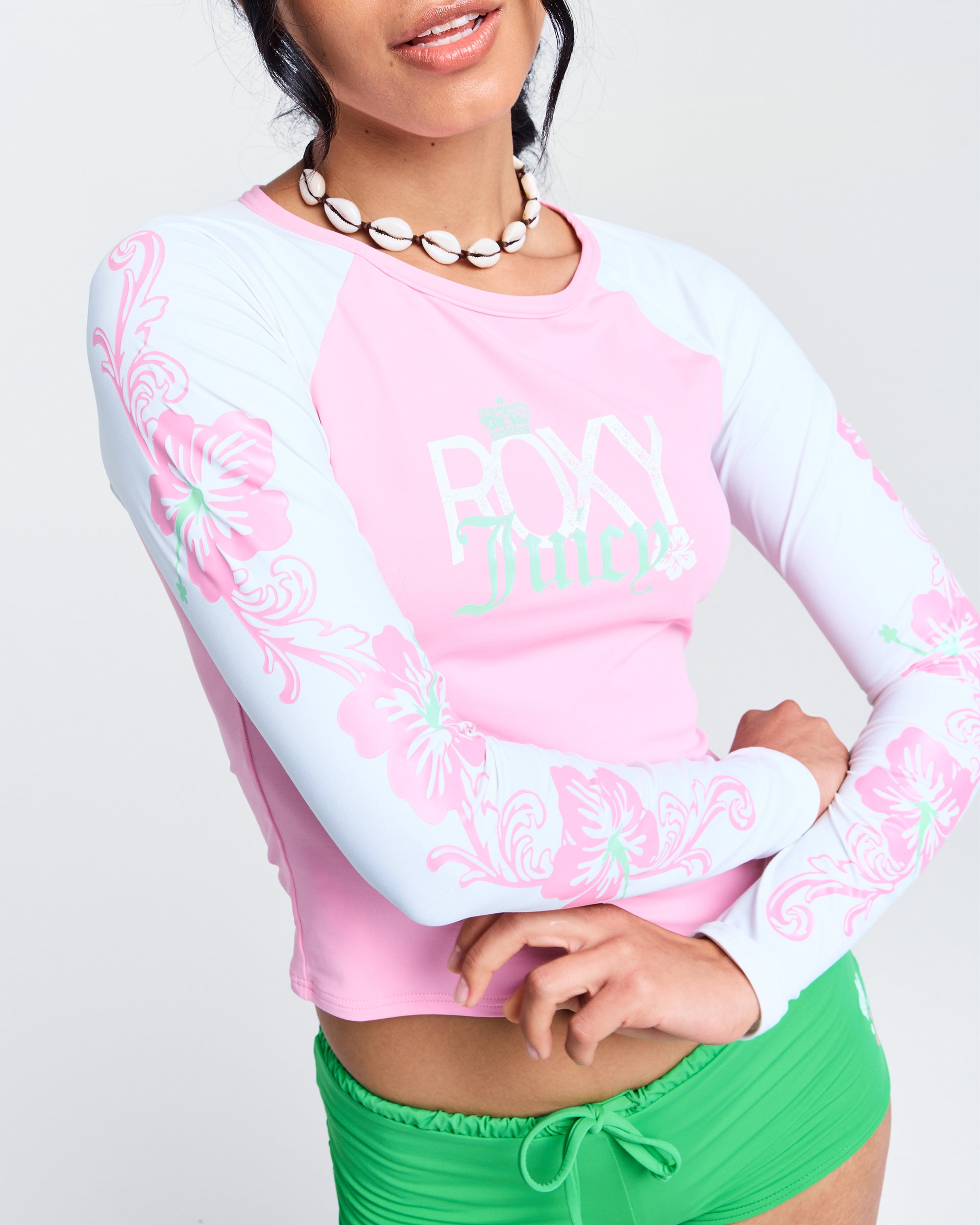 A woman in a pink and white Roxy x Juicy Long Sleeve Lycra Rashguard with floral designs, green swim bottoms, and a shell necklace poses with her arms crossed and lips slightly parted.