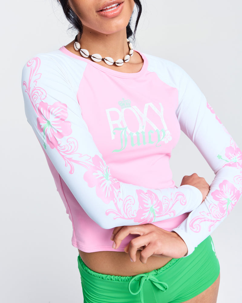 A woman in a pink and white Roxy x Juicy Long Sleeve Lycra Rashguard with floral designs, green swim bottoms, and a shell necklace poses with her arms crossed and lips slightly parted.