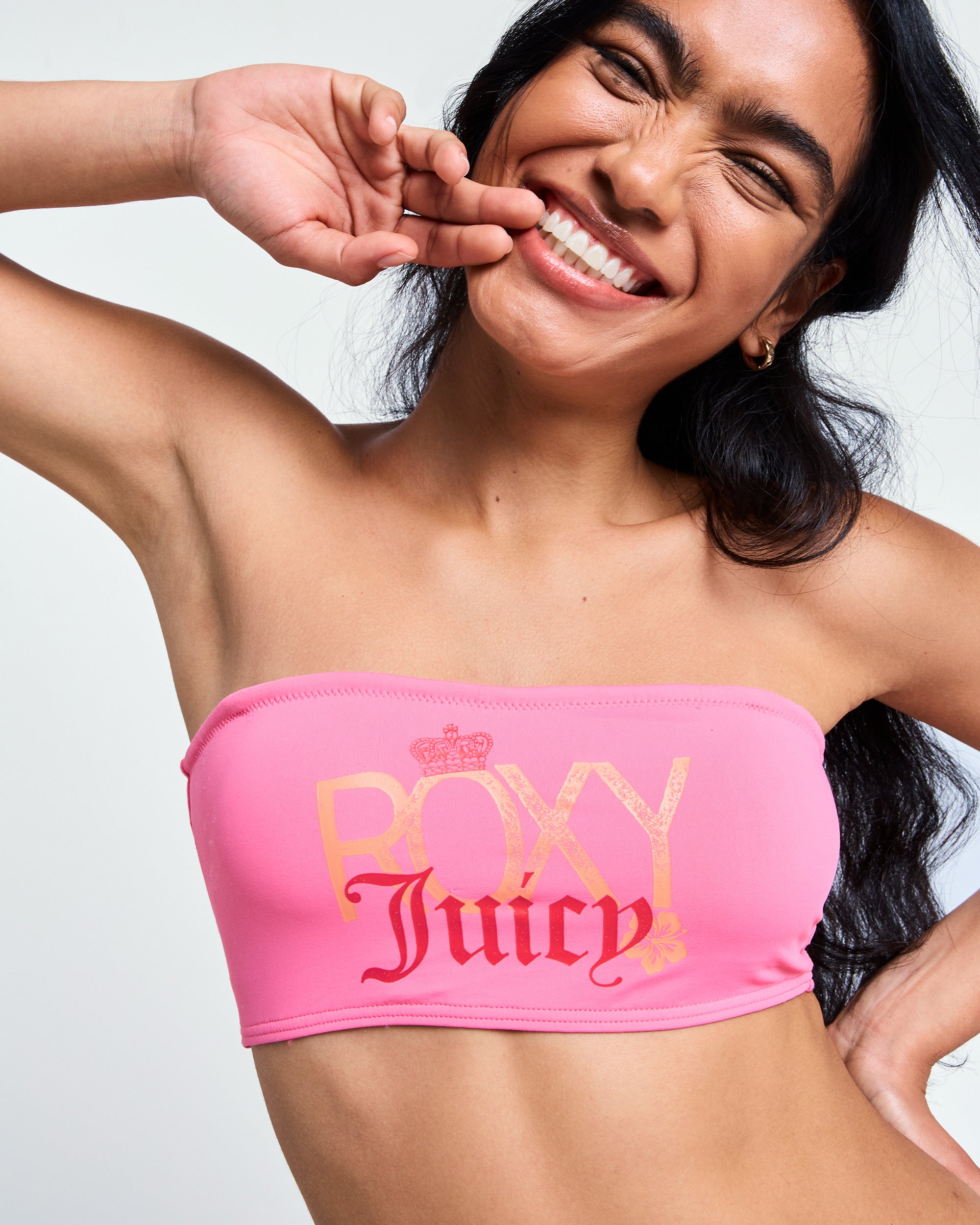 A woman with long dark hair smiles brightly, holding her hand near her face. She wears the Roxy x Juicy Bandeau Bikini Top by Roxy, crafted from recycled nylon and featuring "ROXY Juicy" in bold yellow and red letters.
